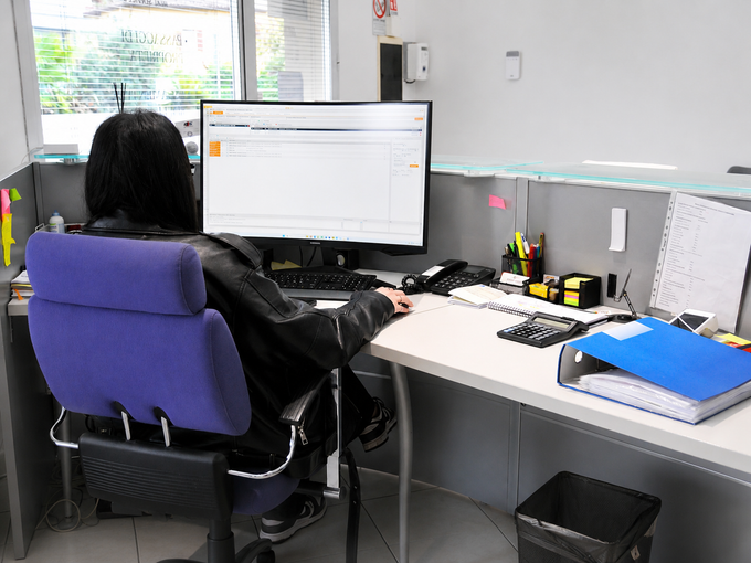 Una persona seduta a una scrivania utilizza un computer in un luminoso cubicolo d'ufficio, con documenti e una cartella blu nelle vicinanze.