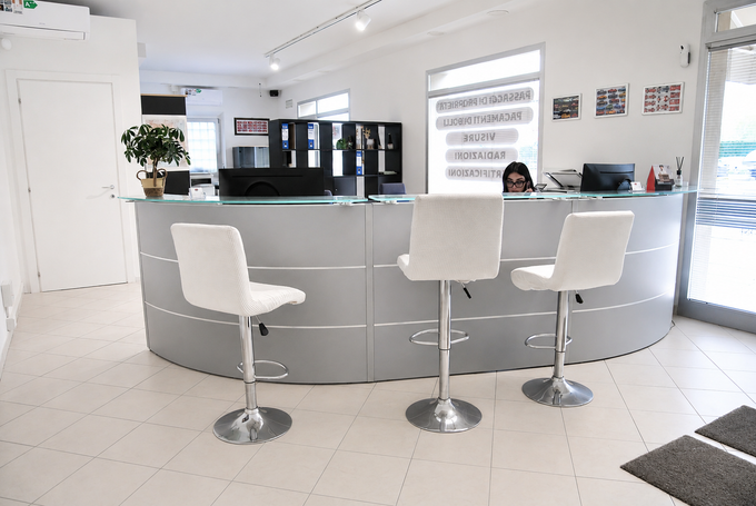 Bancone reception bianco brillante con due sgabelli bianchi in una hall moderna, mensole e una persona sullo sfondo.