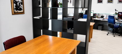 Ufficio con tavolo in legno, pile di documenti, scaffali neri e scrivanie sullo sfondo.