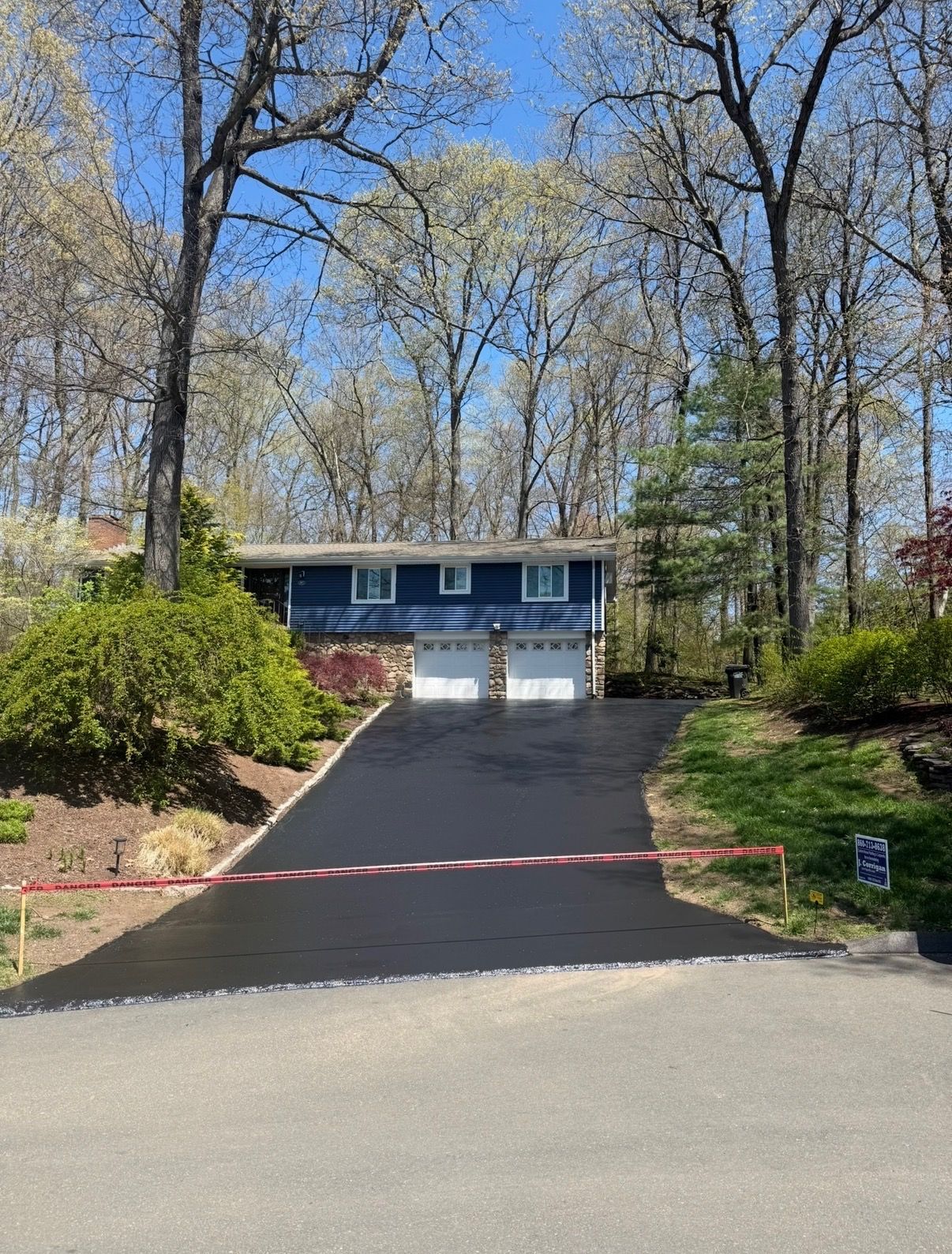 Blue two-story house at the top of a steep driveway, surrounded by trees and shrubs under a clear sky.