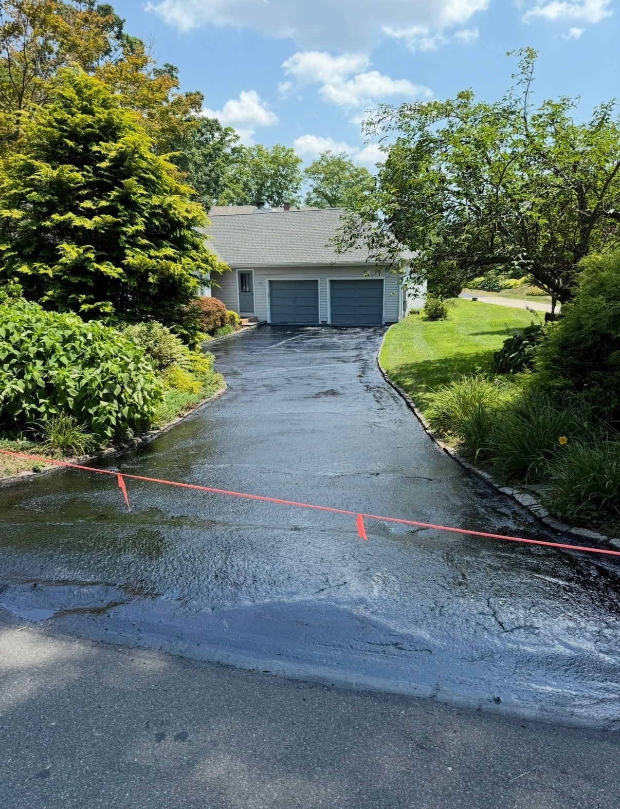 Wet driveway leading to a gray garage, bordered by green trees and grass, with a red tape barrier in front