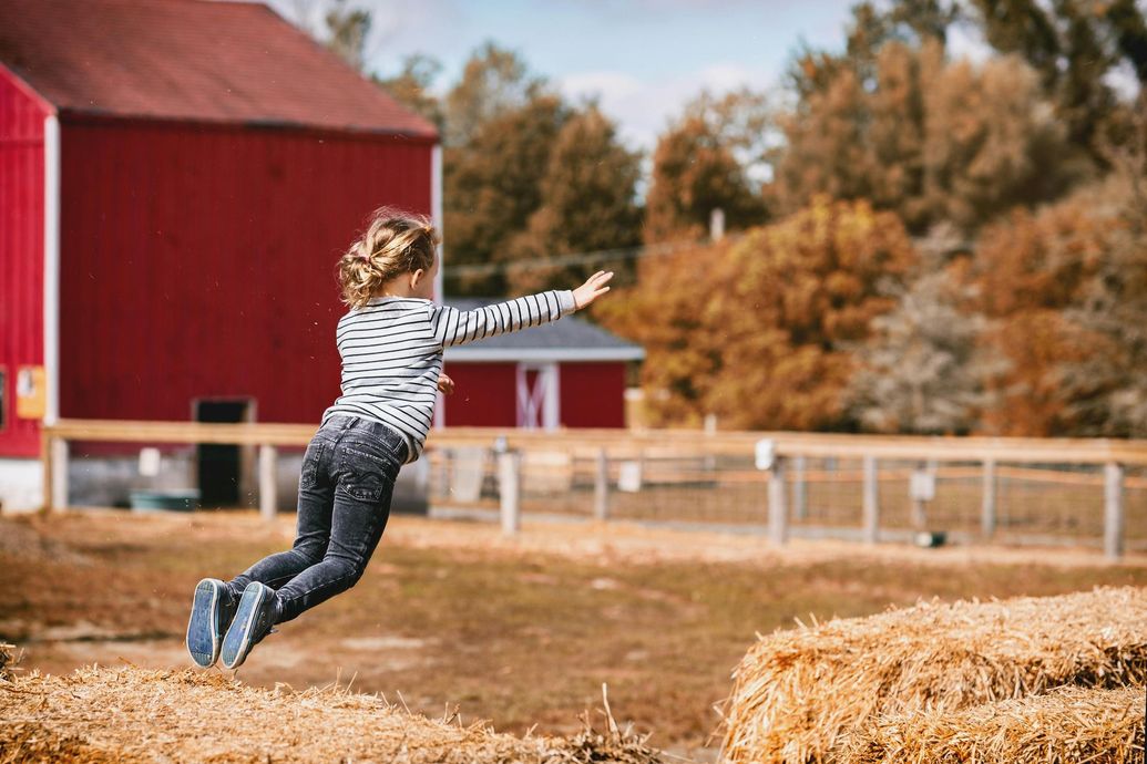 Child jumping toward hay bales near a red barn in a rural field