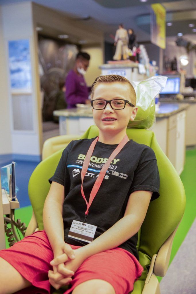 Person sitting in a green chair at an indoor event, wearing glasses, a black shirt, and pink shorts.