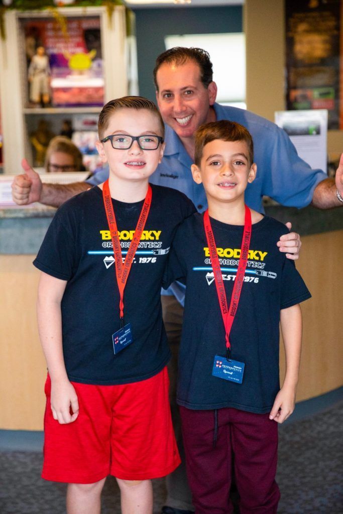 Two boys wearing medals stand with a smiling man in a shop or lobby.