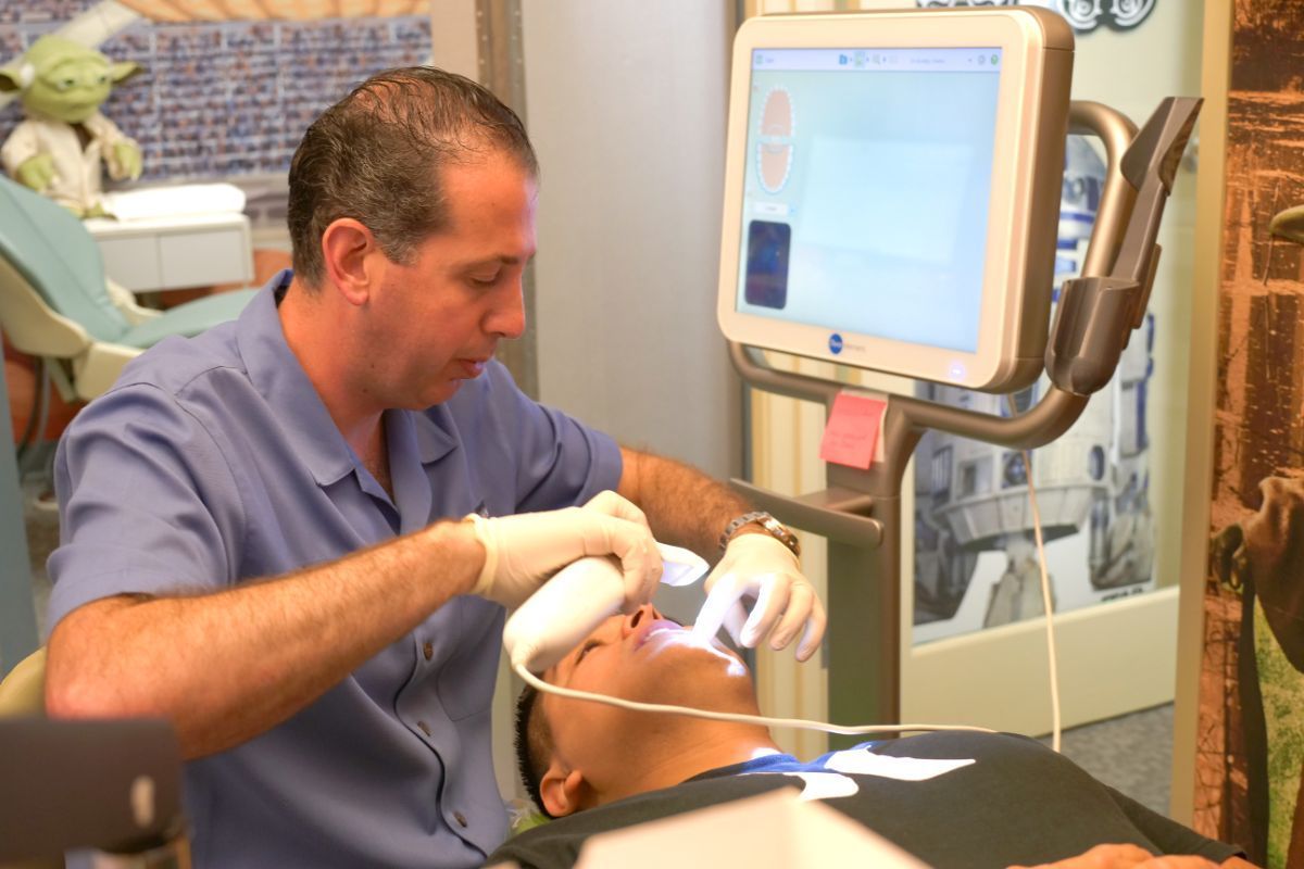 Dentist wearing gloves examines a patient in a dental chair beside a monitor.