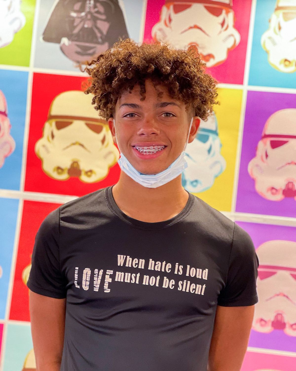 Smiling person in a black shirt standing before a colorful pop-art wall of masked faces