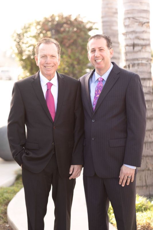 Two men in dark suits and pink ties standing outdoors by palm trees.