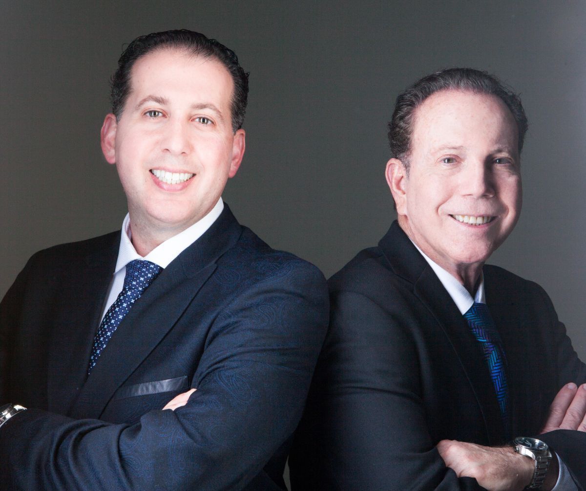 Two men in dark suits smiling with arms crossed against a gray studio background