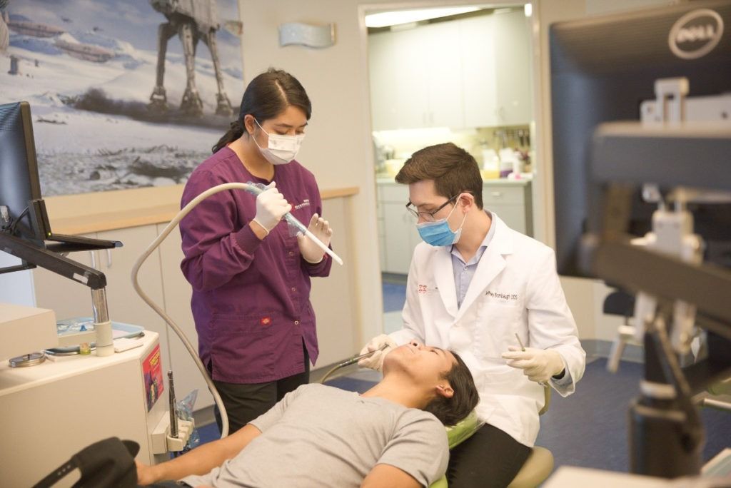 Dentist treating a patient while a masked assistant stands nearby in a clinic room