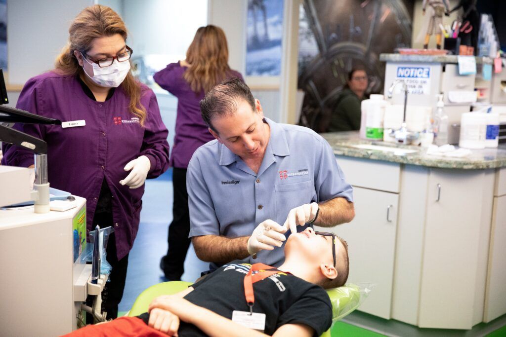 Dentist treating a patient in a chair while a masked assistant stands nearby in a clinic room