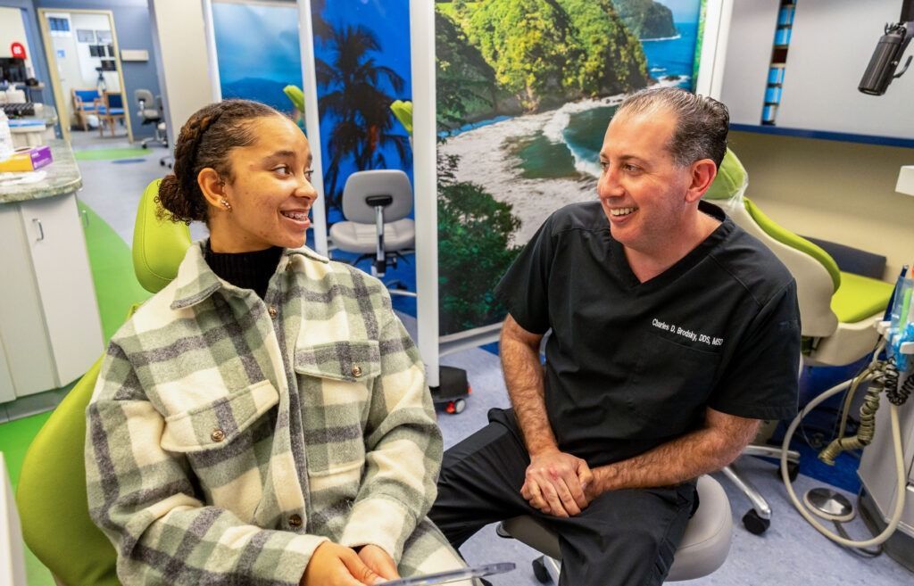 Two people smiling in hospital chairs, with medical equipment and a window view behind them.