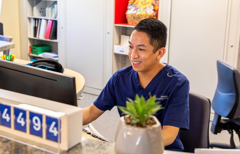 Smiling person at a desk in an office, with computer monitors and a potted plant in front.