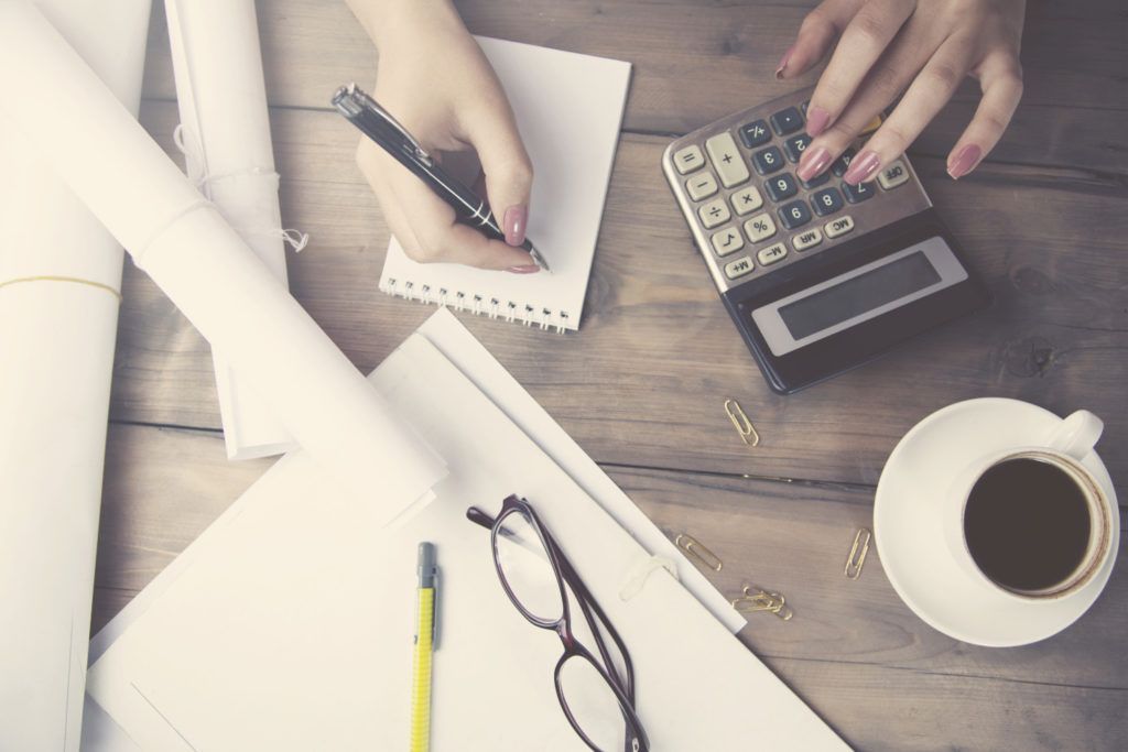Desk with hands using a calculator and writing on paper beside coffee, glasses, and rolled blueprints.