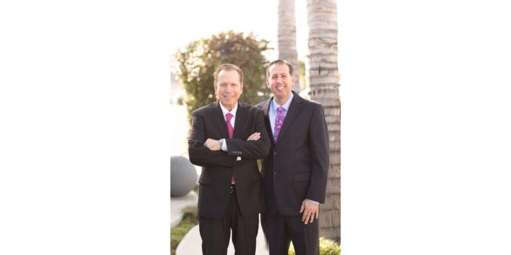 Two men in suits standing outdoors in front of a stone building, one with arms crossed.