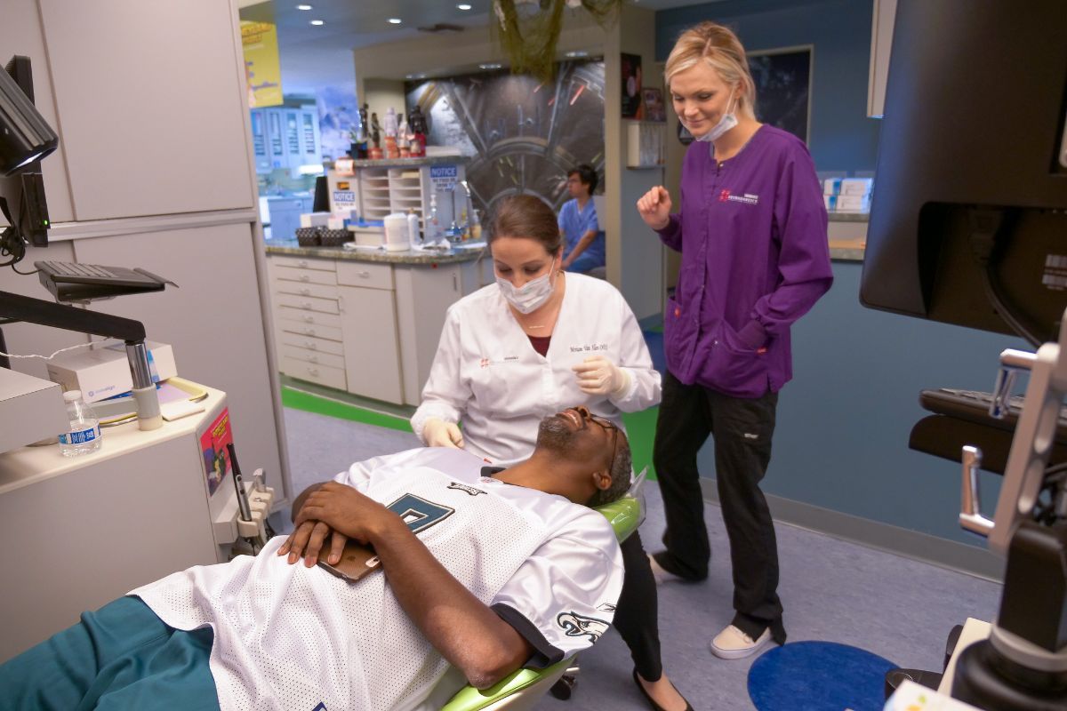 Doctor checks a reclining patient while another clinician stands nearby in a clinic room