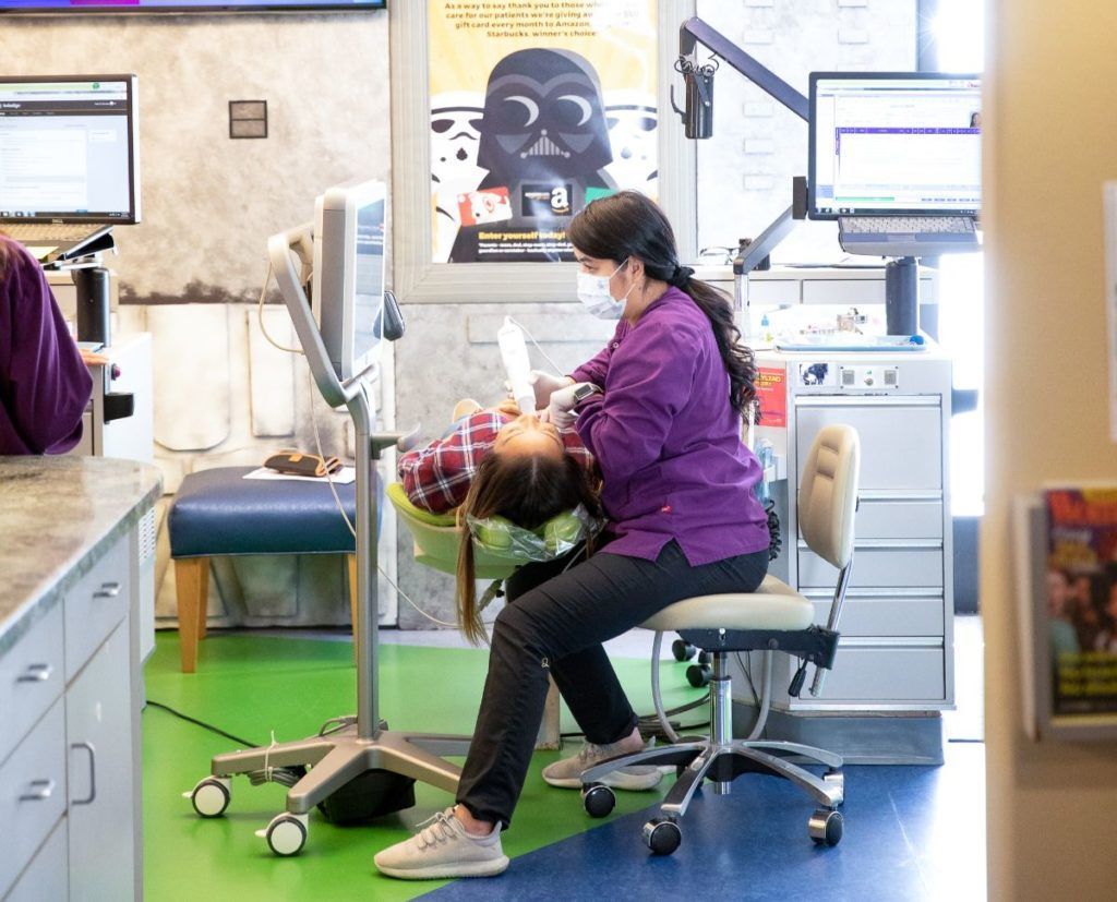 Dentist in purple jacket treating patient in a dental clinic chair, with equipment nearby.