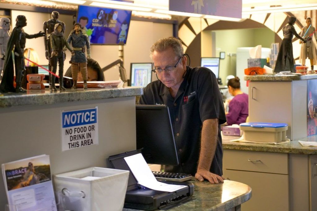 Man leaning over a laptop at a shop counter with shelves, figurines, and a notice sign behind him