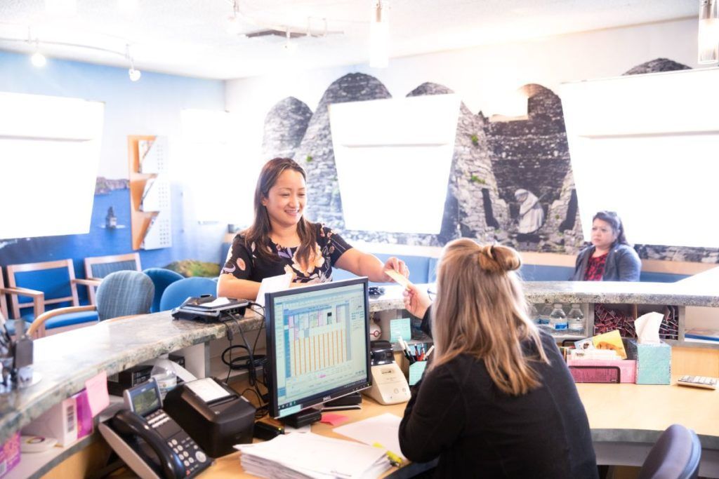 People at a reception desk in a bright office, with computers and paperwork.
