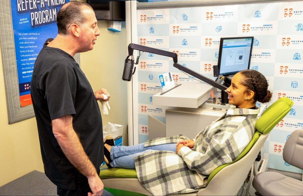 Man speaking with a patient reclined in a green chair in a dental clinic exam room