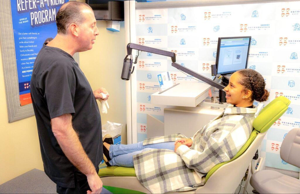 Woman in plaid hospital gown reclining in a clinic chair while a man stands beside her near a monitor.