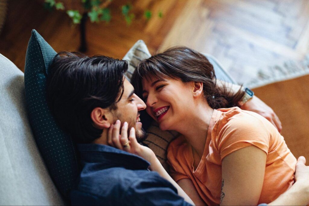 Two people lying close on a couch, smiling warmly at each other in a cozy room.