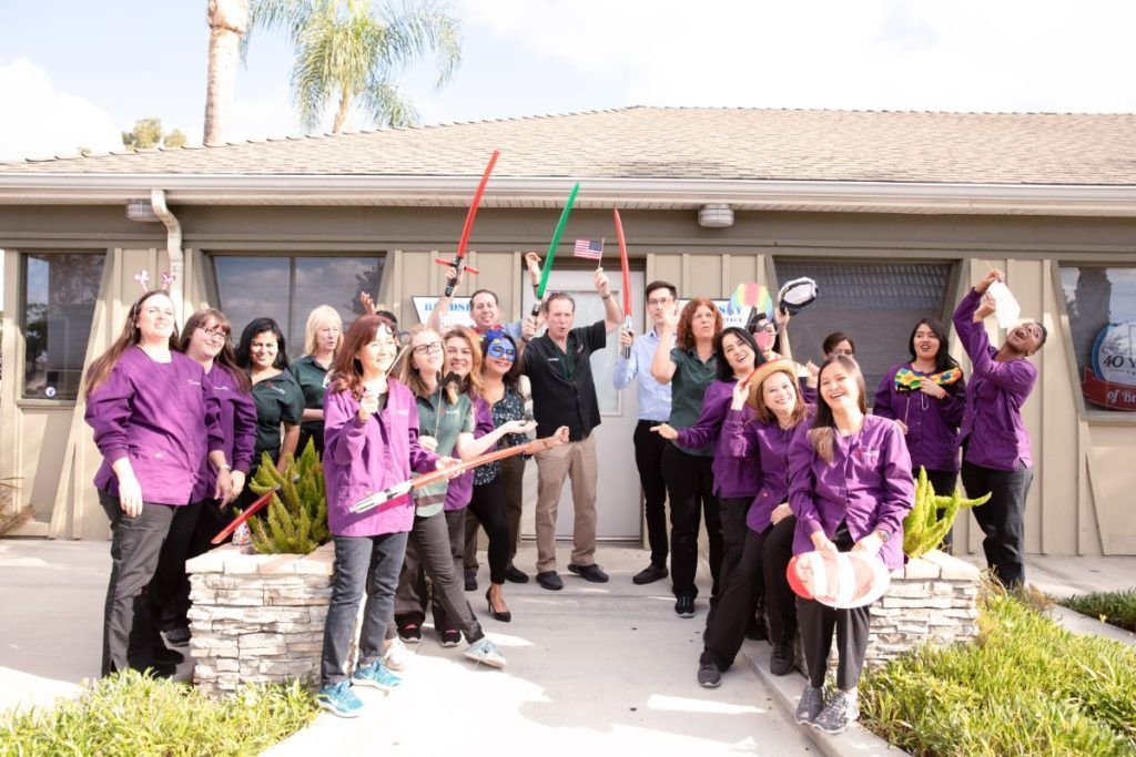 Group in purple shirts posing and cheering outside a house, holding colorful props.