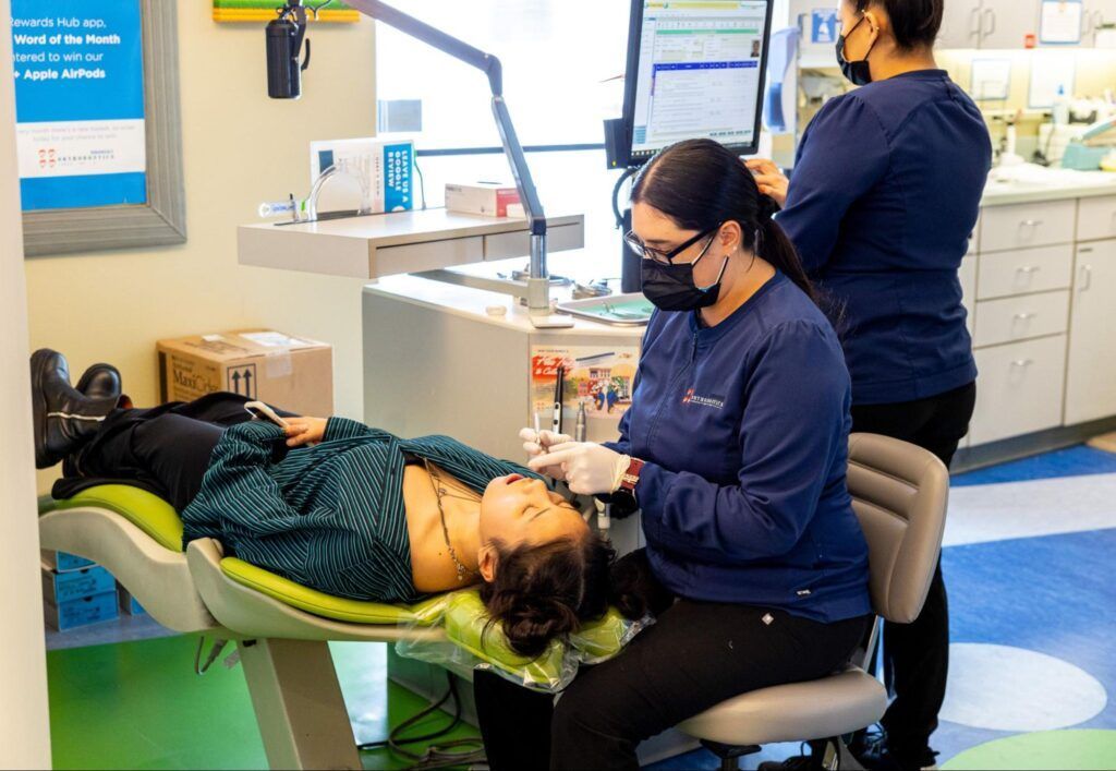 Two masked people in a clinic, one seated while working on a patient lying on an exam chair