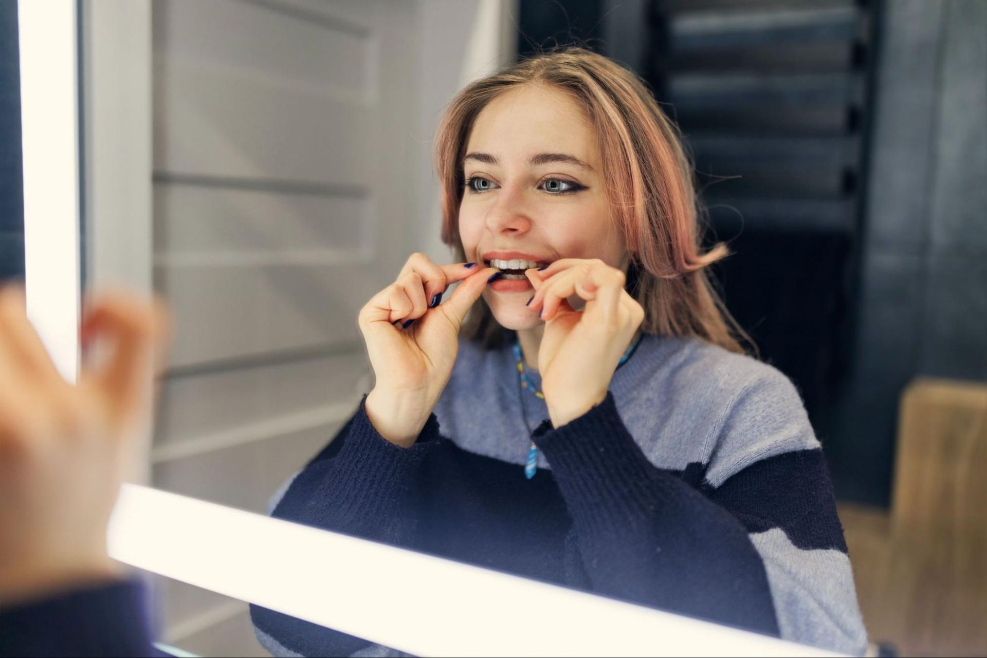 Woman adjusting a mouthguard in front of a mirror, wearing a navy and gray sweater.