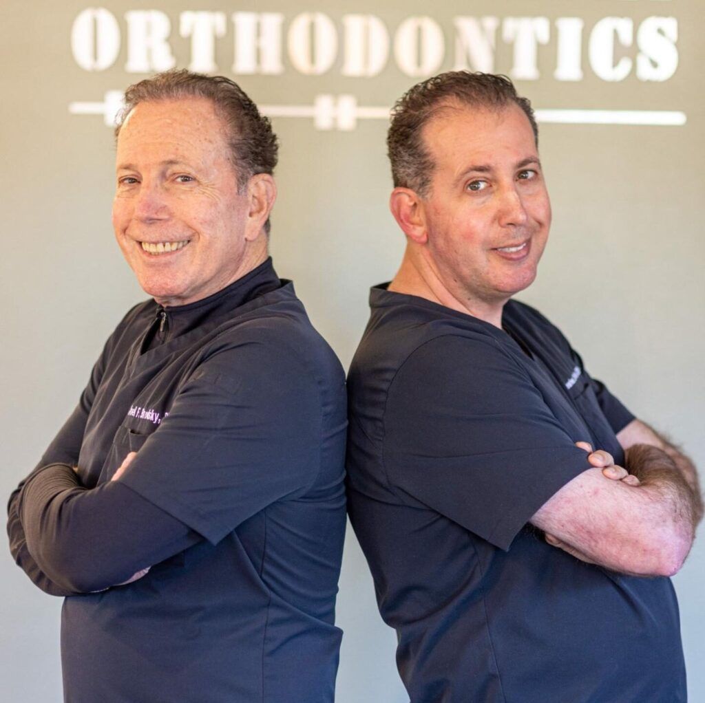 Man in black scrubs posing front and side in an orthodontics office with a “ORTHODONTICS” sign behind him