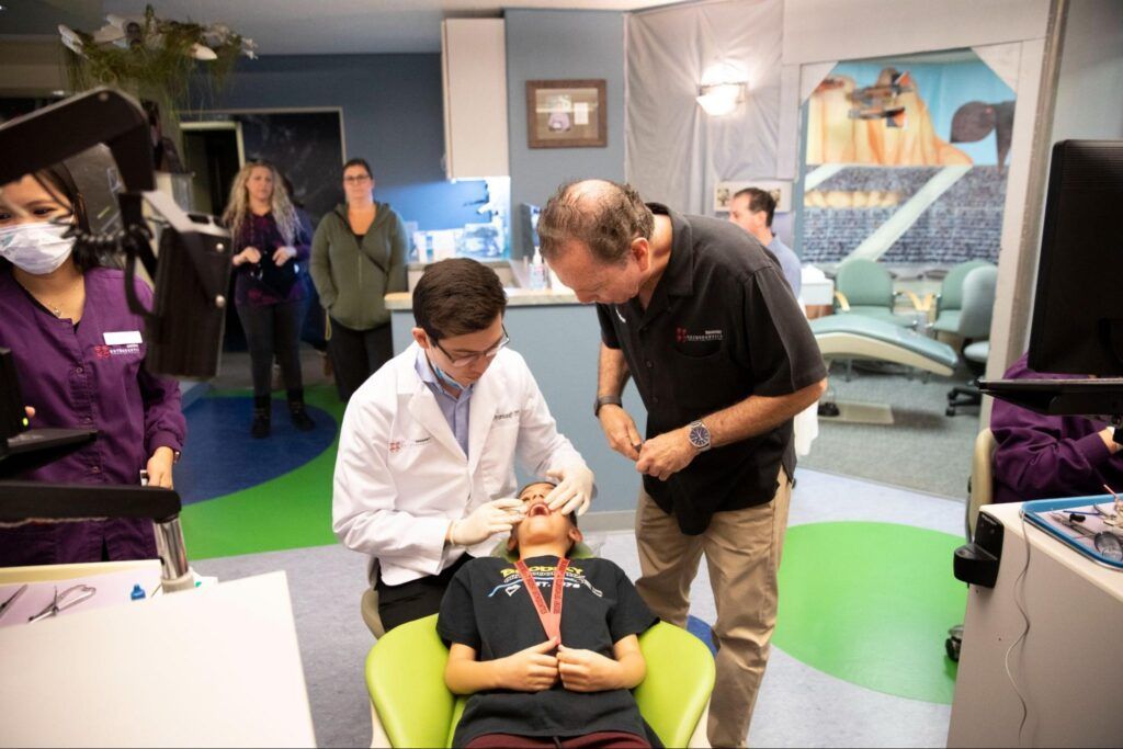 Person in white coat examines seated patient while another man leans in, in a clinic exhibit room