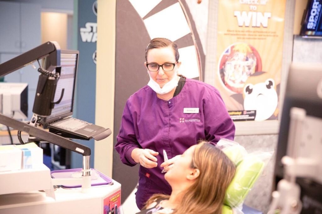 Dentist in purple coat examining a patient in a dental clinic, with a monitor and equipment nearby.