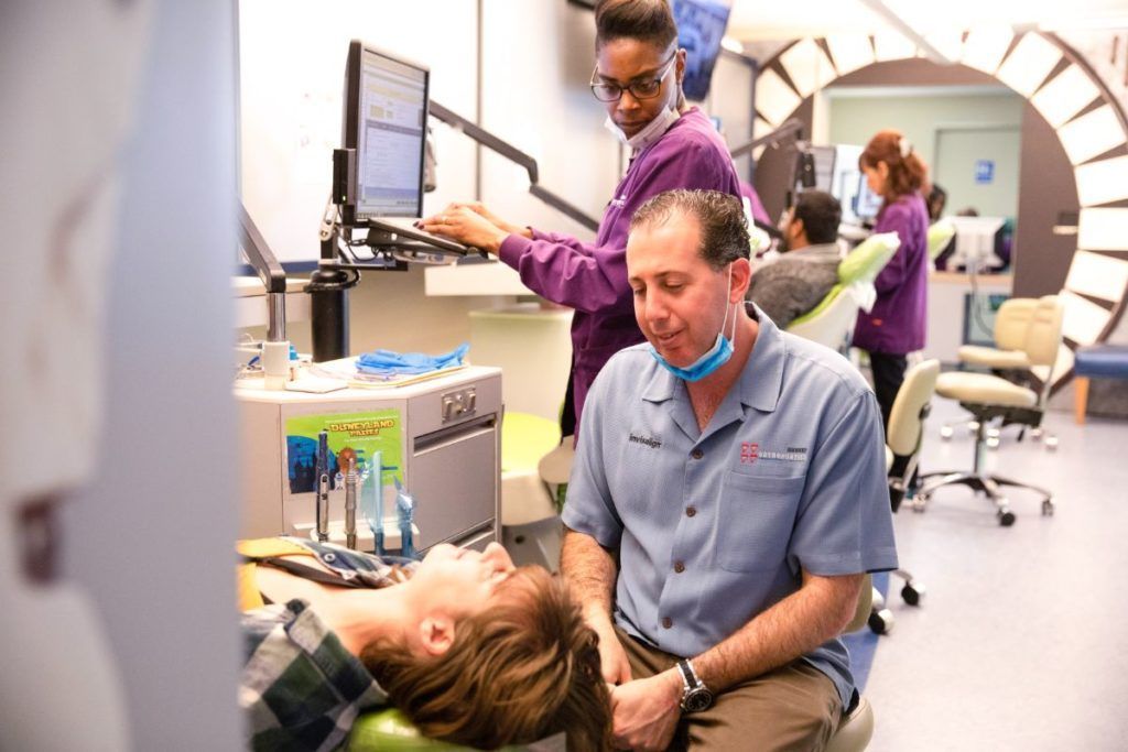 Dental clinic with patient in chair, dentist examining her, and assistant preparing equipment nearby