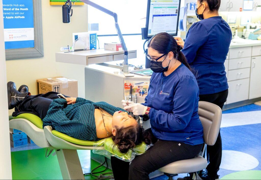 Dentist treating a patient in a bright clinic, with another staff member nearby.