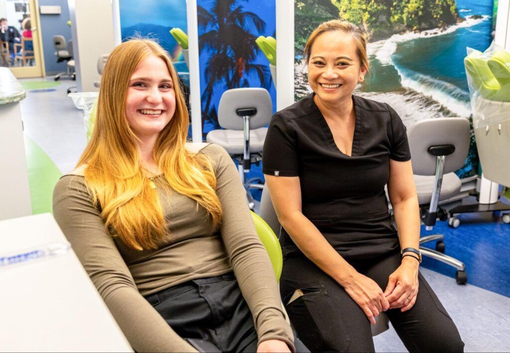 Two people sitting in office chairs in a bright room with a beach mural, smiling at the camera