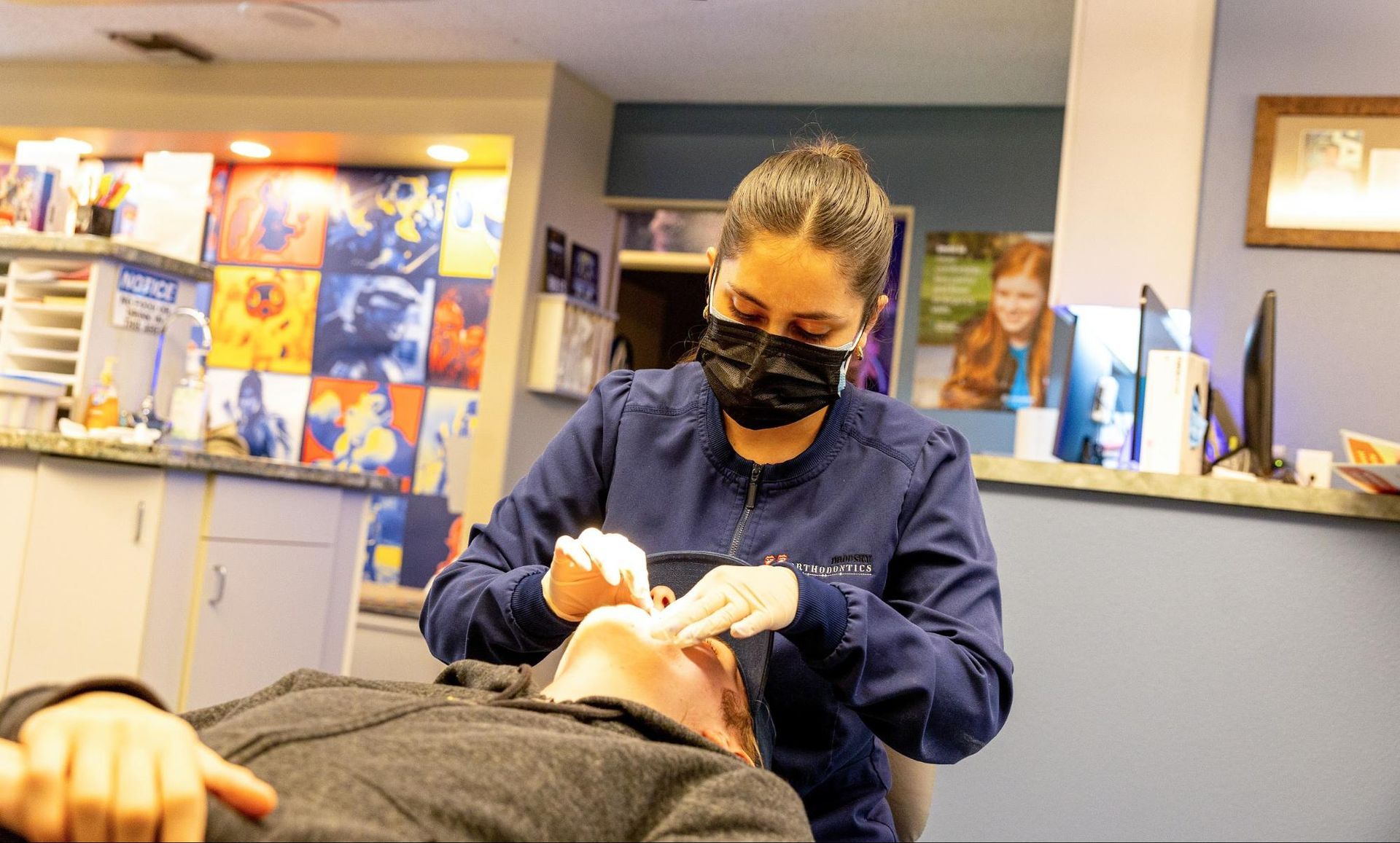 Dental hygienist wearing a mask examines a patient in a clinic, with posters and a reception desk behind.