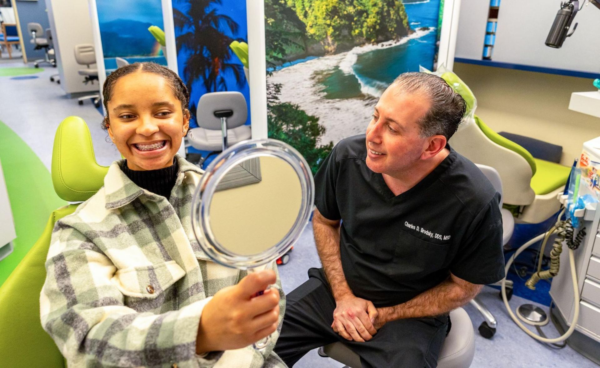 Woman smiling into handheld mirror while seated in a dental office, with a man beside her.