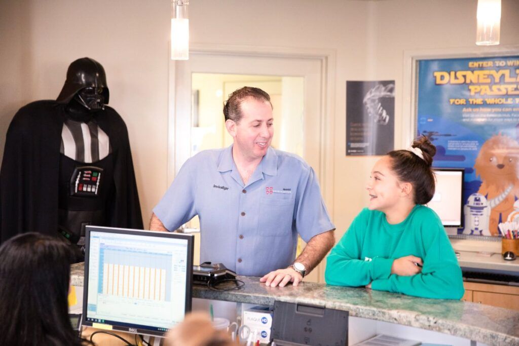 Two people talk at a counter near a Darth Vader statue in a bright lobby.