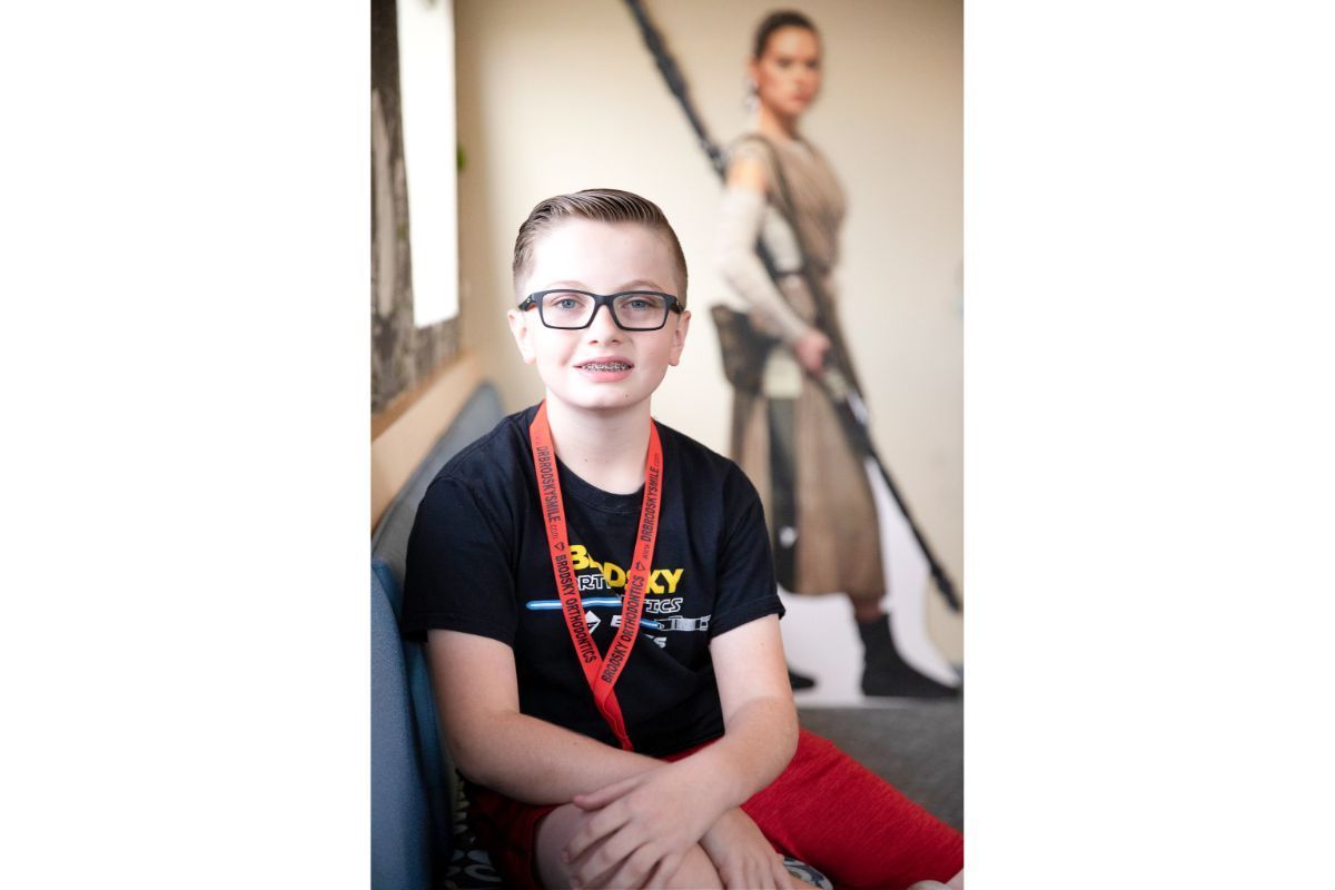 Person wearing glasses and a lanyard sits indoors, with a blurred figure standing in the background.