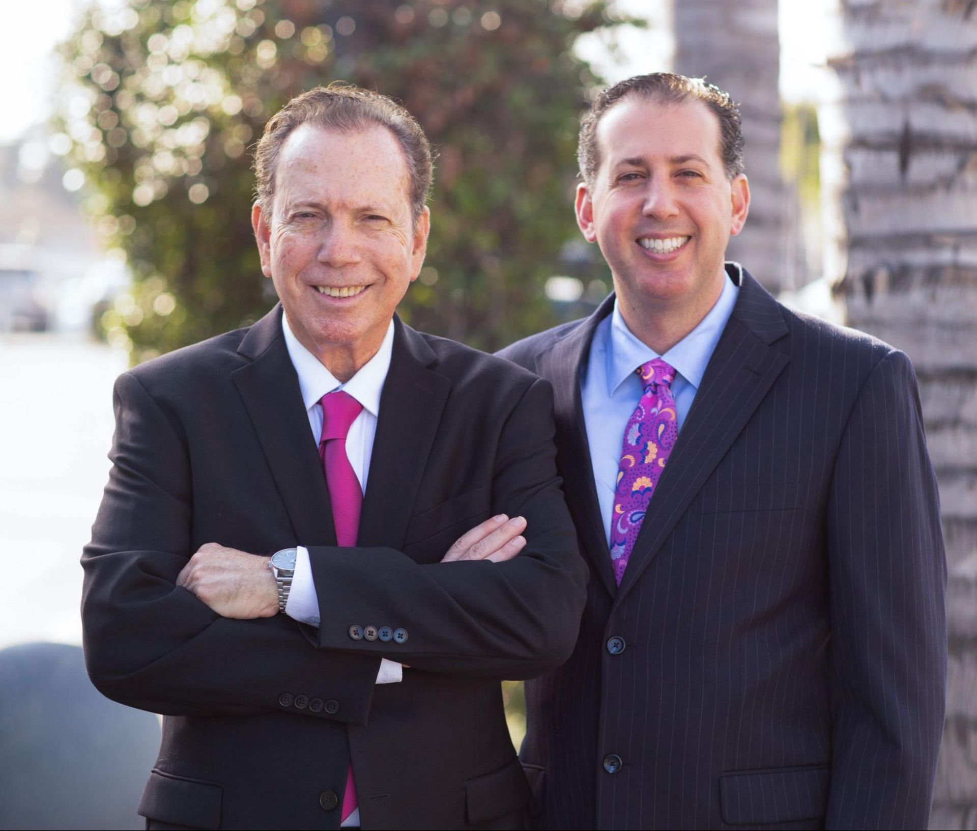 Two men in dark suits standing outdoors, one with arms crossed, both smiling.