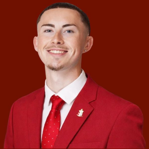 Man in red suit and tie against a red backdrop. He has a serious expression and a lapel pin.