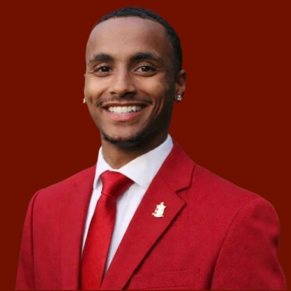 Young Black man in a red suit and tie, smiling, against a burgundy background.