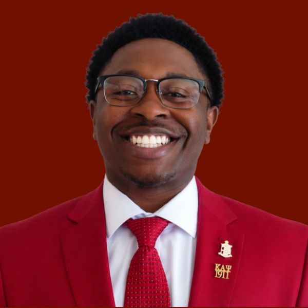 Young Black man in a red suit and tie, professional headshot with a burgundy background.