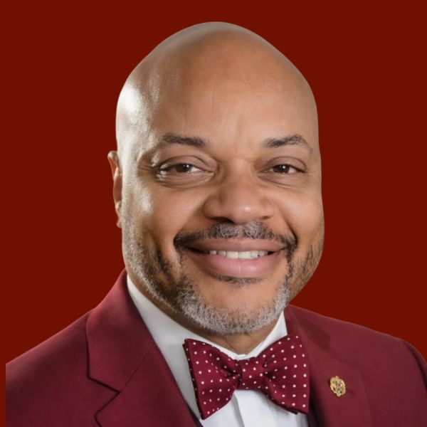 Man in red suit smiles at the camera, maroon tie and red background.