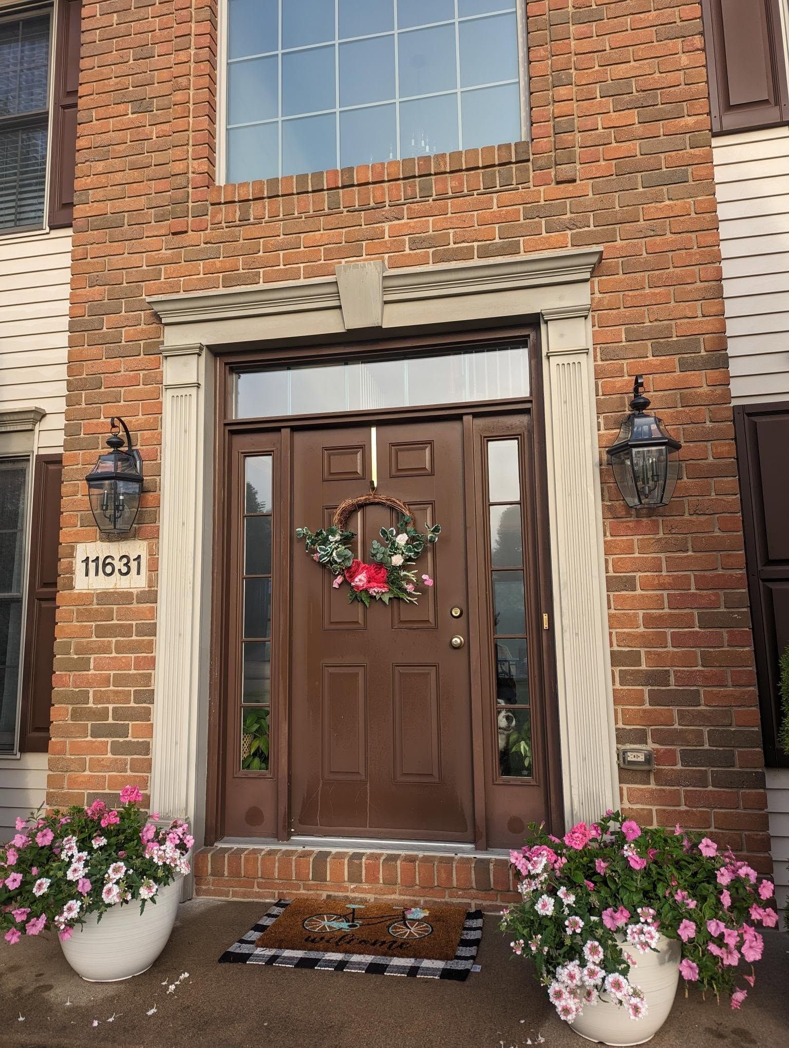 Brick house entrance with brown double doors, holiday wreath, lanterns, and pink potted flowers.