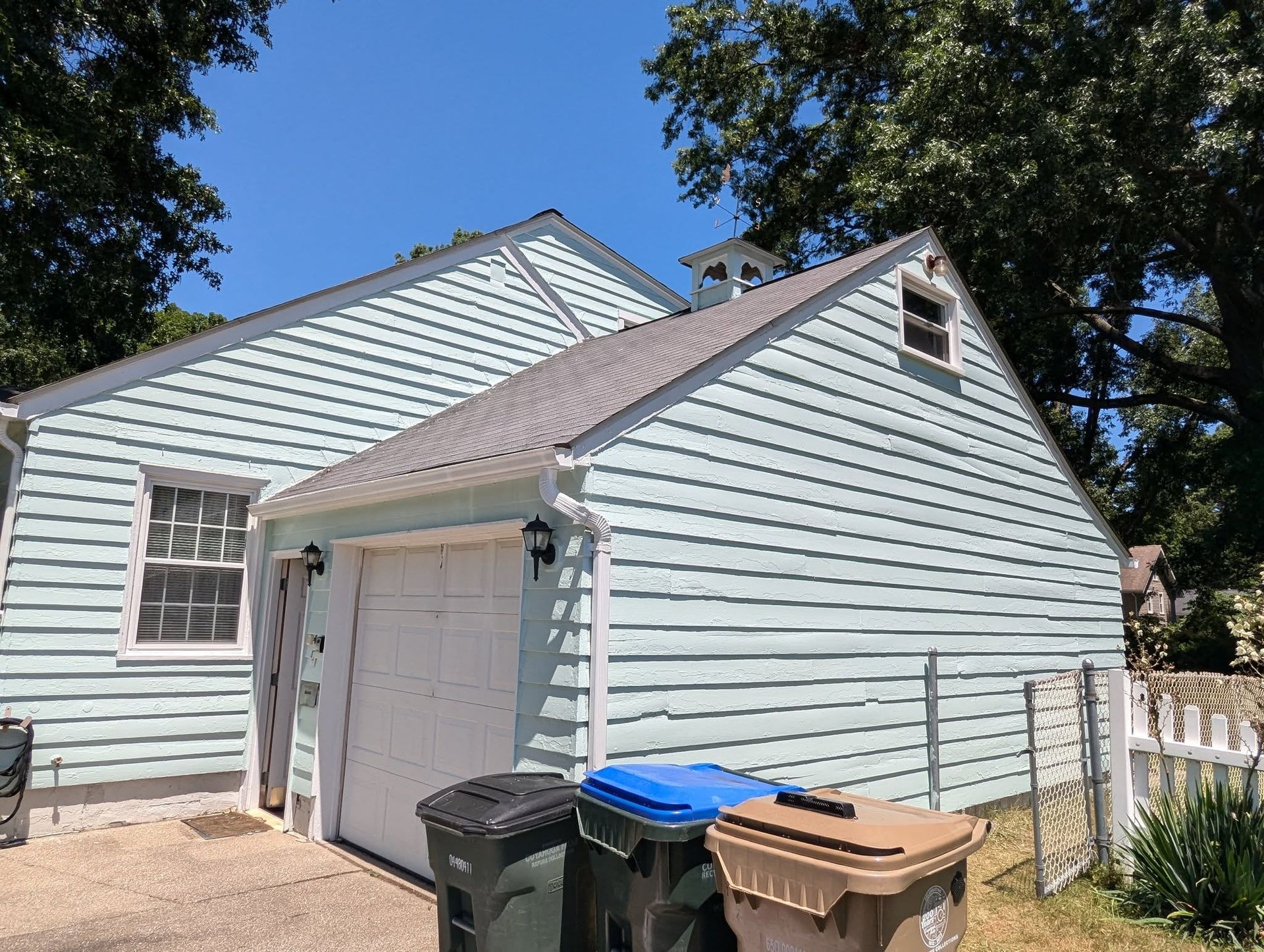 Light blue house exterior with attached garage, trash bins, and trees in the yard