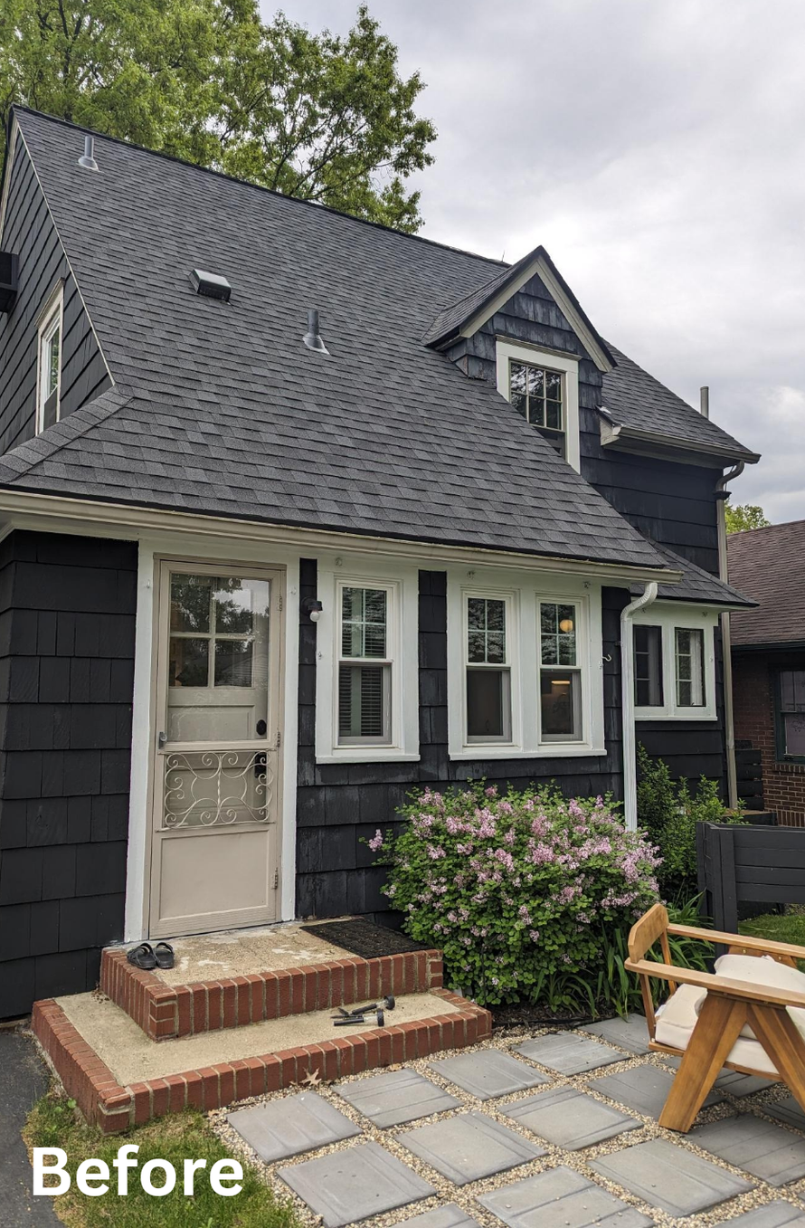 Small dark-shingled house exterior with white-trimmed windows, front door, brick steps, and patio tiles
