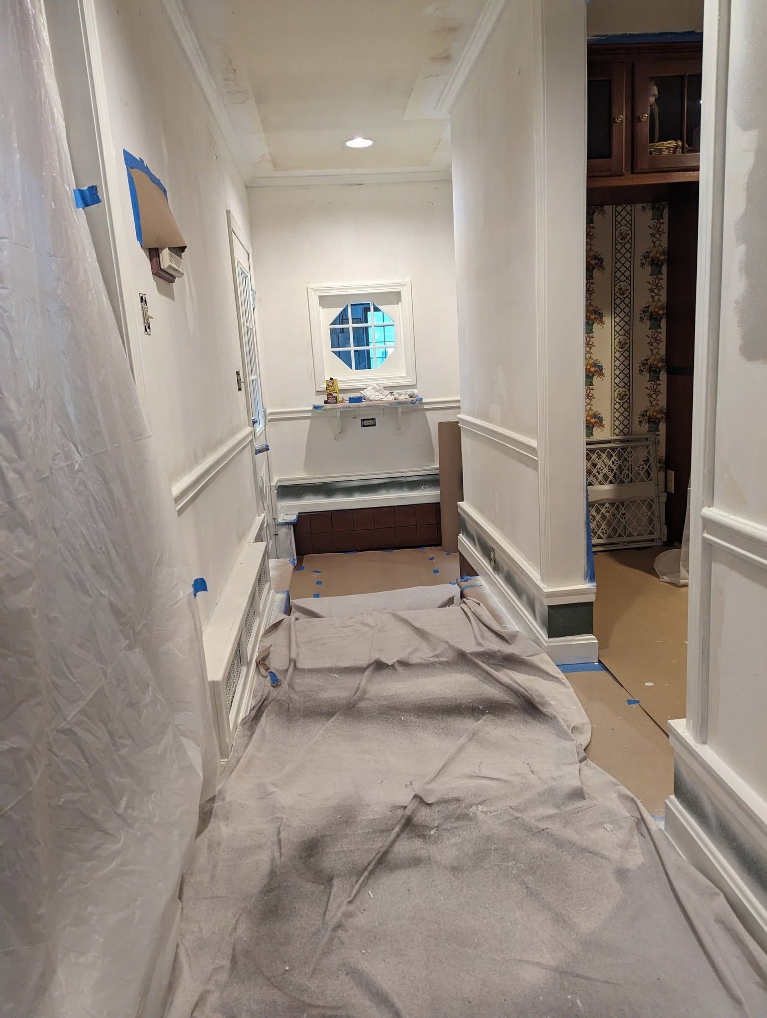 Narrow hallway under renovation, with covered floors, taped walls, and a small window at the end.
