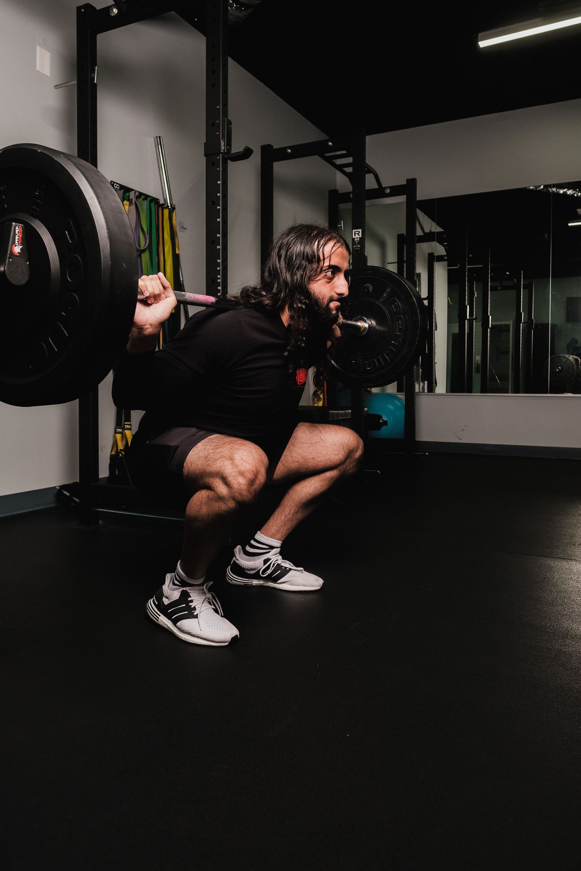 Person squats with a barbell in a dim gym, wearing black workout clothes and sneakers.