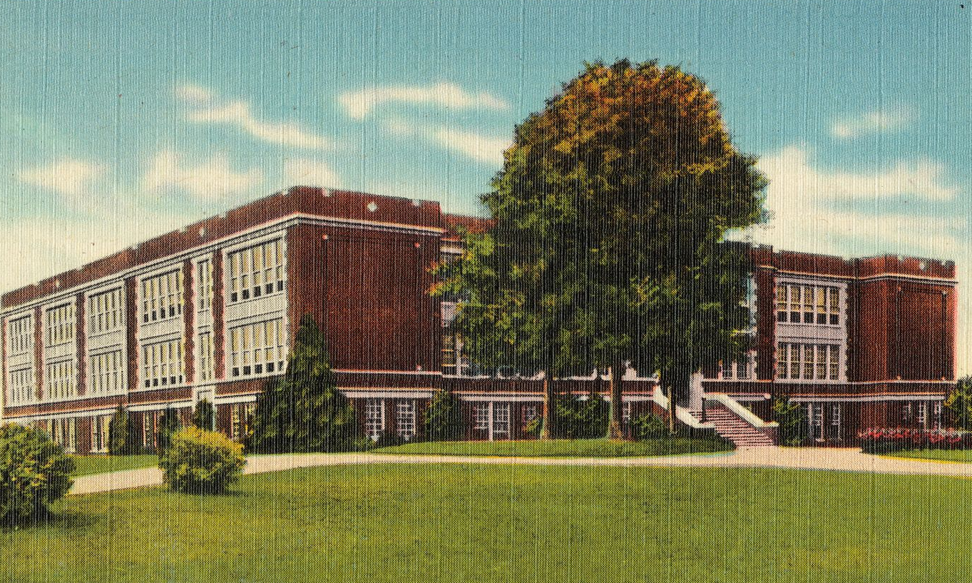 Red brick school building with many windows, trees, and a grassy lawn.