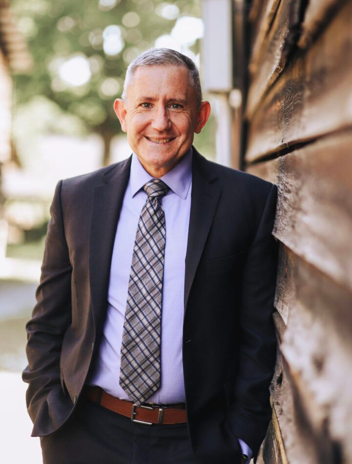 Man in suit leans against a wooden wall, smiling. He wears a tie and a purple shirt.